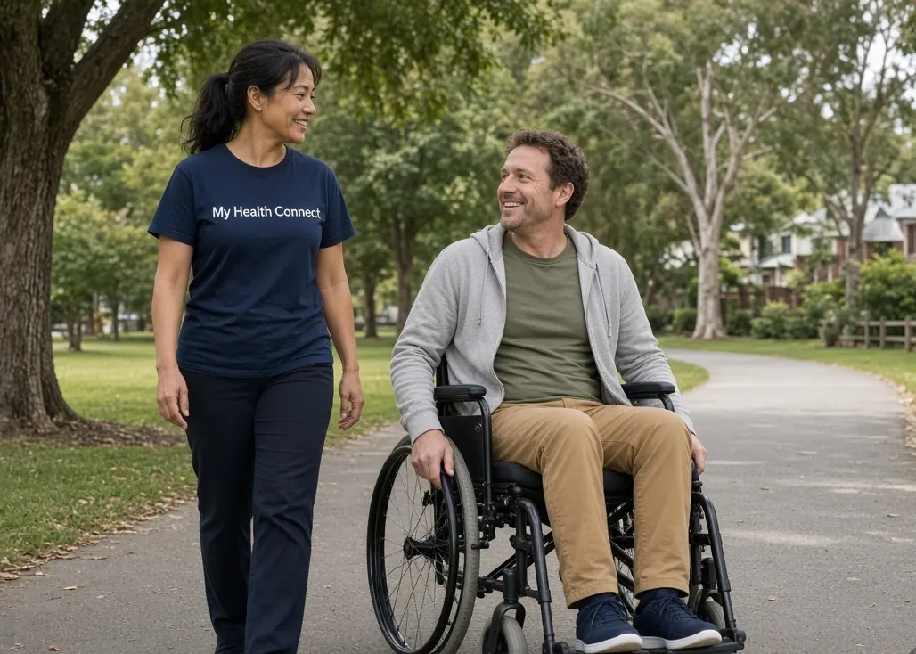A My Health Connect support worker walking alongside a participant in his wheelchair through a leafy park.